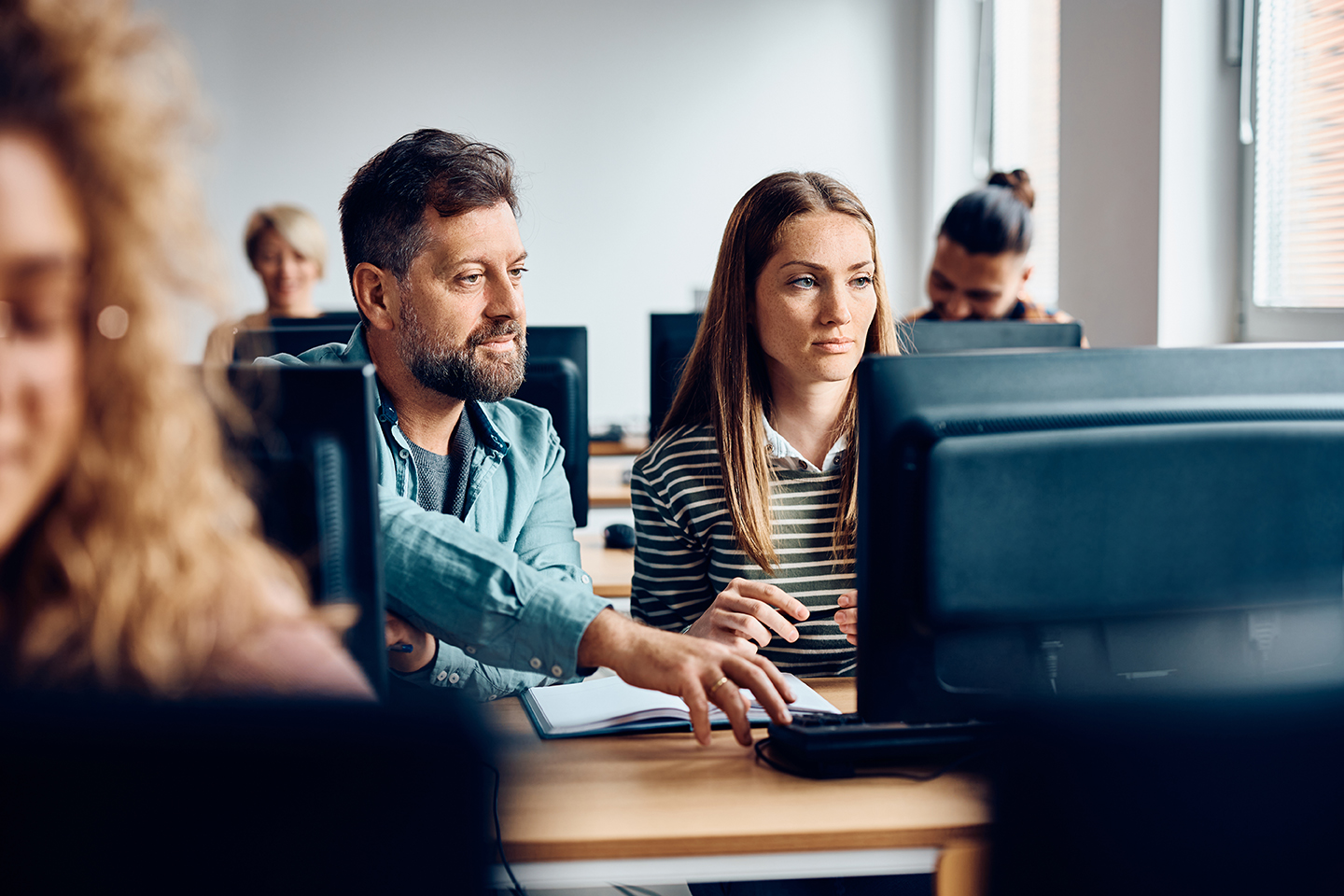 Adult students cooperating while e-learning on a computer in the classroom. Mid adult woman and her mature classmate using desktop PC during computer class at the university.